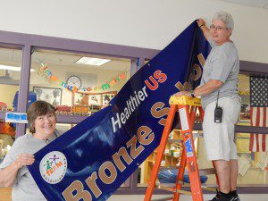 Gateway’s Food Services Director Wendy McCaul, left, gets a hand from Littleville Custodian Joanne Frappier to hang the school’s “Healthier U.S. Schools” banner.  (Photo submitted)