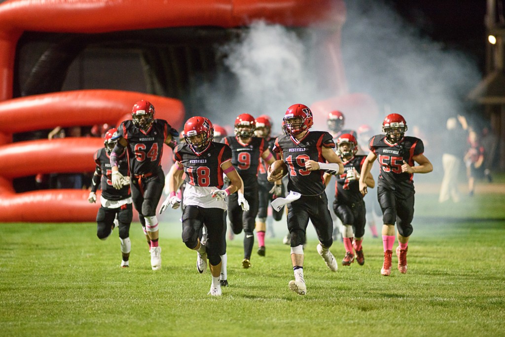 The 2016 Westfield High School football team runs out of a "tunnel" of smoke in a large inflatable helmet to begin a regular season game at Bullens Field. (Photo by Marc St. Onge)